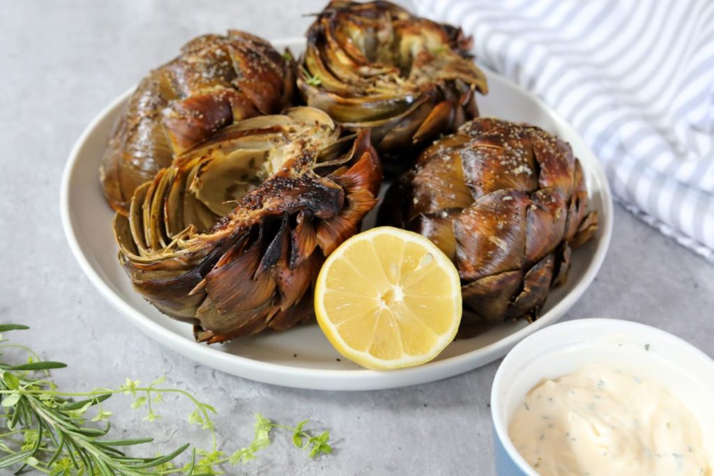 Roasted artichokes on a white plate with a ramekin filled with herbed aioli