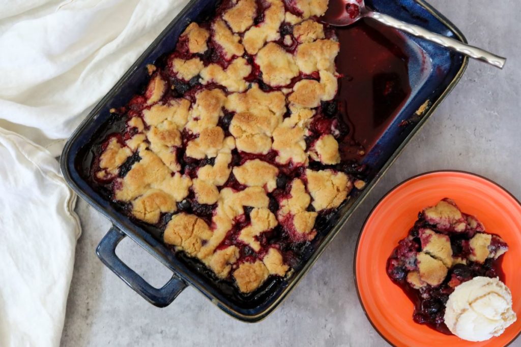 berry cobbler in a baking dish with a serving on an orange plate with a scoop of ice cream