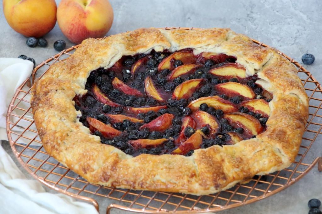 blueberry peach galette on a rose colored cooling rack surrounded by peaches and blueberries