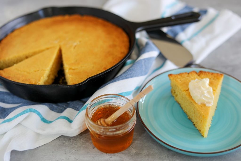 Buttered wedge of cornbread on a blue plate with a cup of honey next to it.