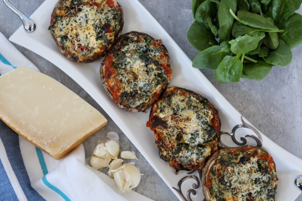 View of all four stuffed portobello mushrooms on a white and sliver tray with spinach, garlic, and a large block of parmesan cheese
