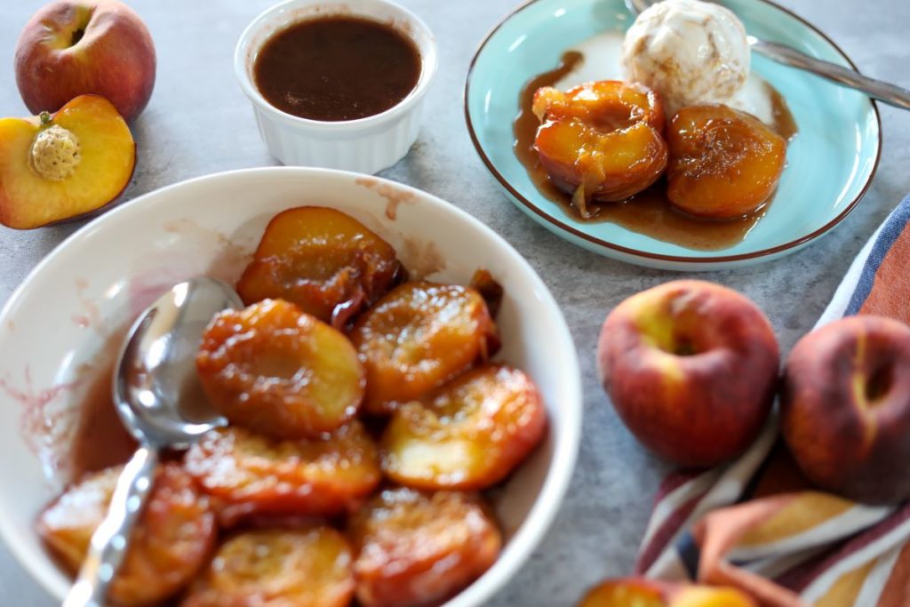 A bowl of bourbon baked peaches with extra sauce on the side with a blue plate with 2 peach halves and ice cream on it.