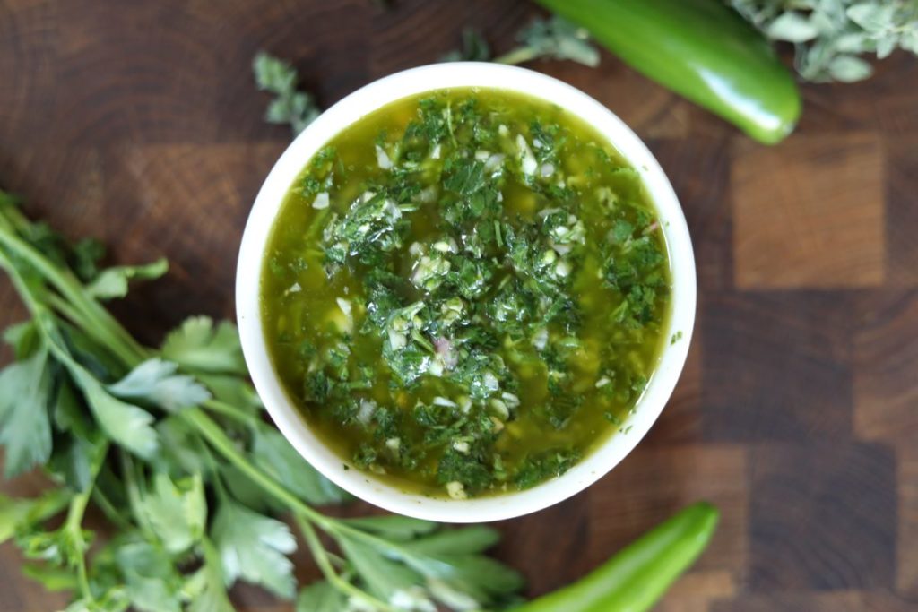 Bowl of chimichurri on a wood cutting board surrounded by some of the ingredients