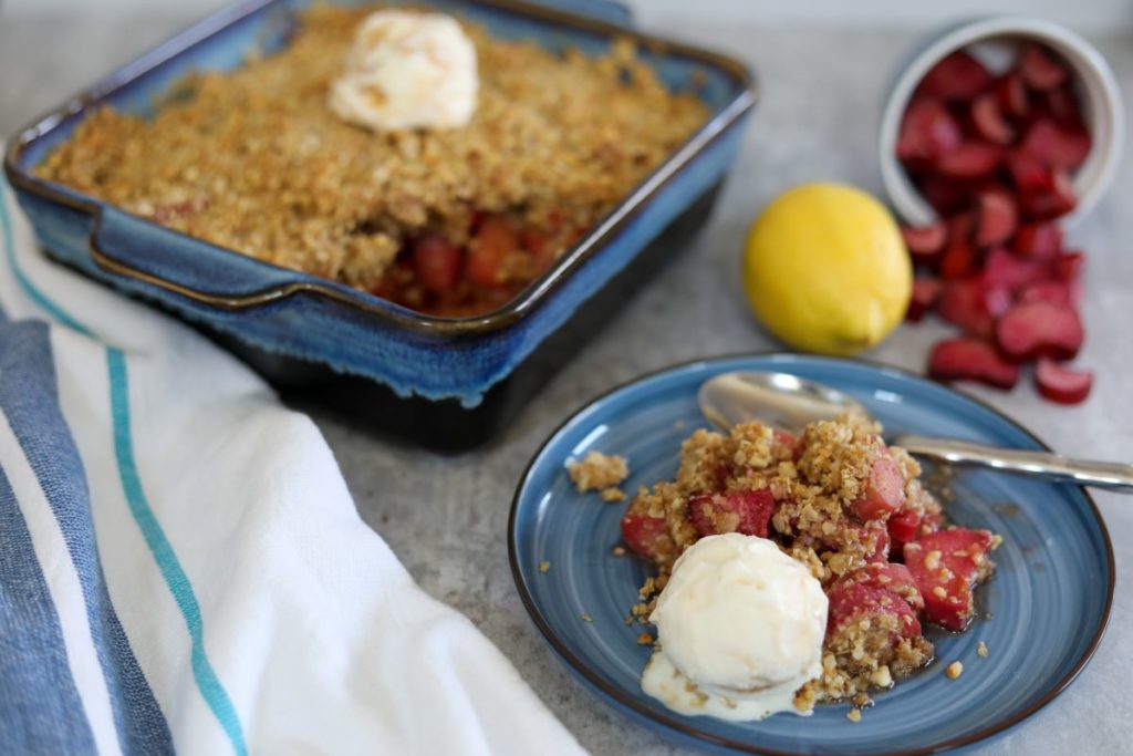 Rhubarb crisp on a blue plate with the baking dish full of the remaining crisp topped with ice cream.