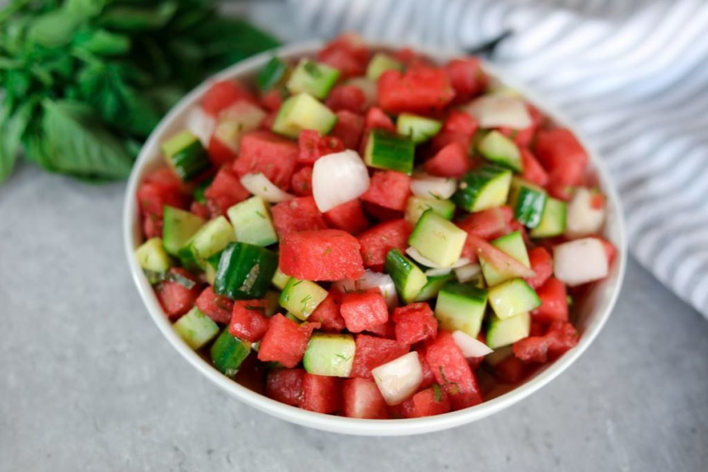 Watermelon salad in a while bowl in from of a grey striped towel and fresh basil