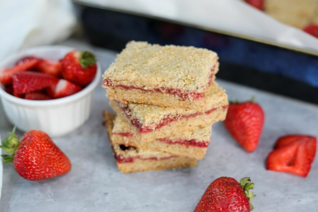 strawberry crumble bars stacked next to a bowl of fresh strawberries