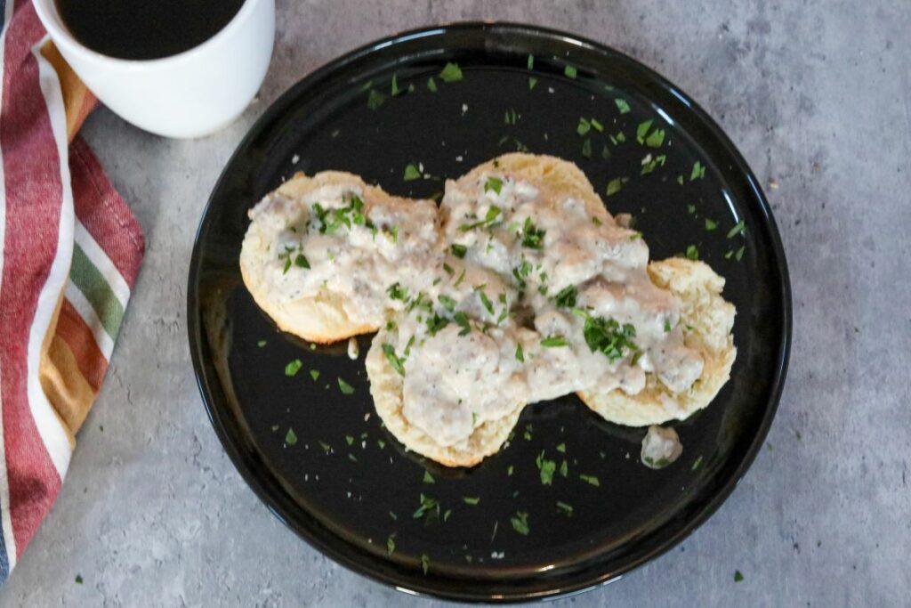 Biscuits and gravy on a black plate next to a cup of coffee