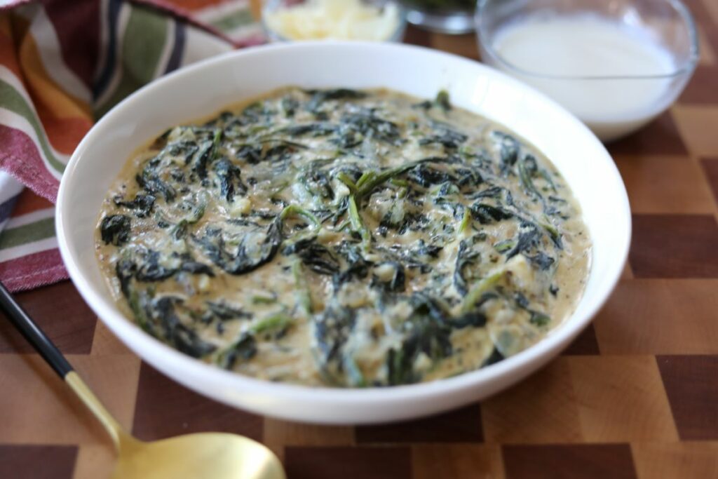 A white bowl of creamed spinach with a gold serving spoon