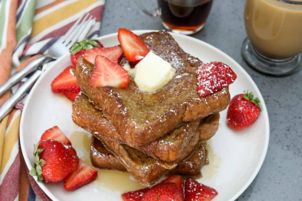A stack of French toast on a white plate with strawberries and syrup
