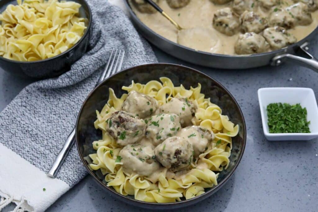 A bowl of Swedish meatballs on egg noodles with a pan of the remaining meatballs.