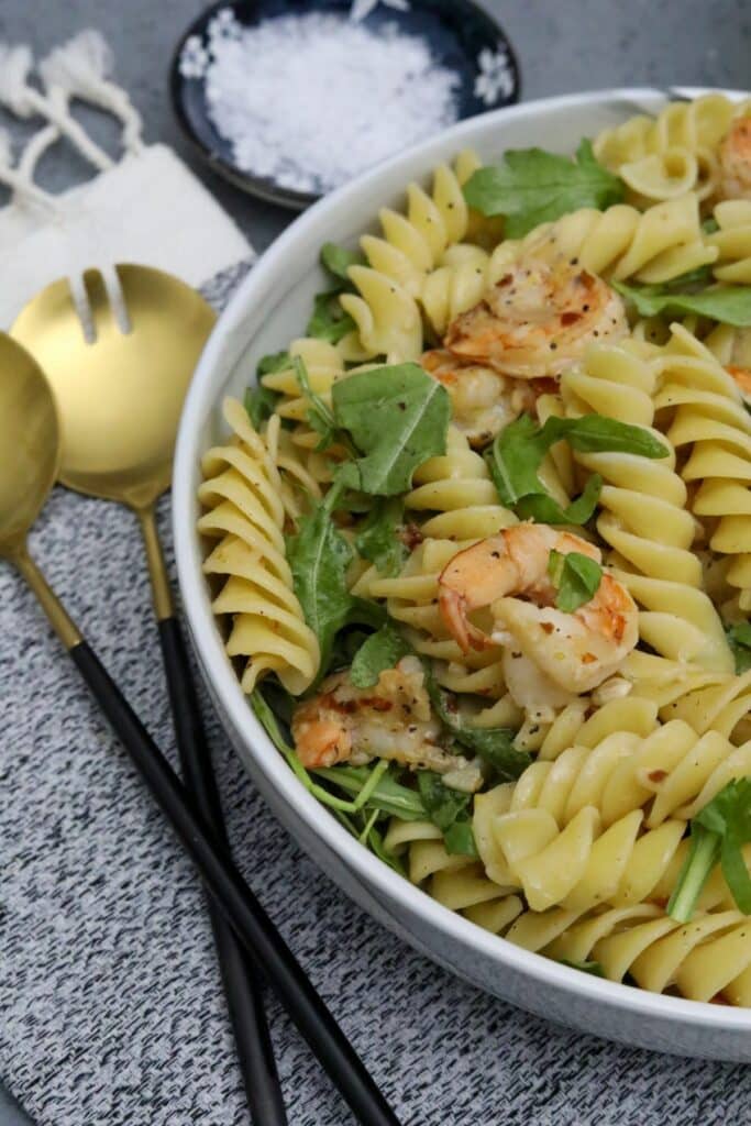 A bowl with shrimp fusilli and arugula with a serving utensils on a cloth napkin