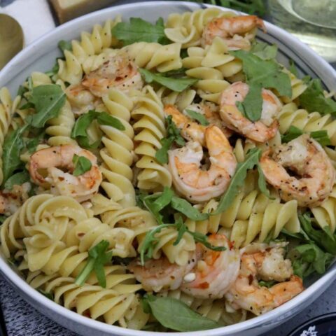 A serving bowl with shrimp fusilli and arugula with a glass of wine